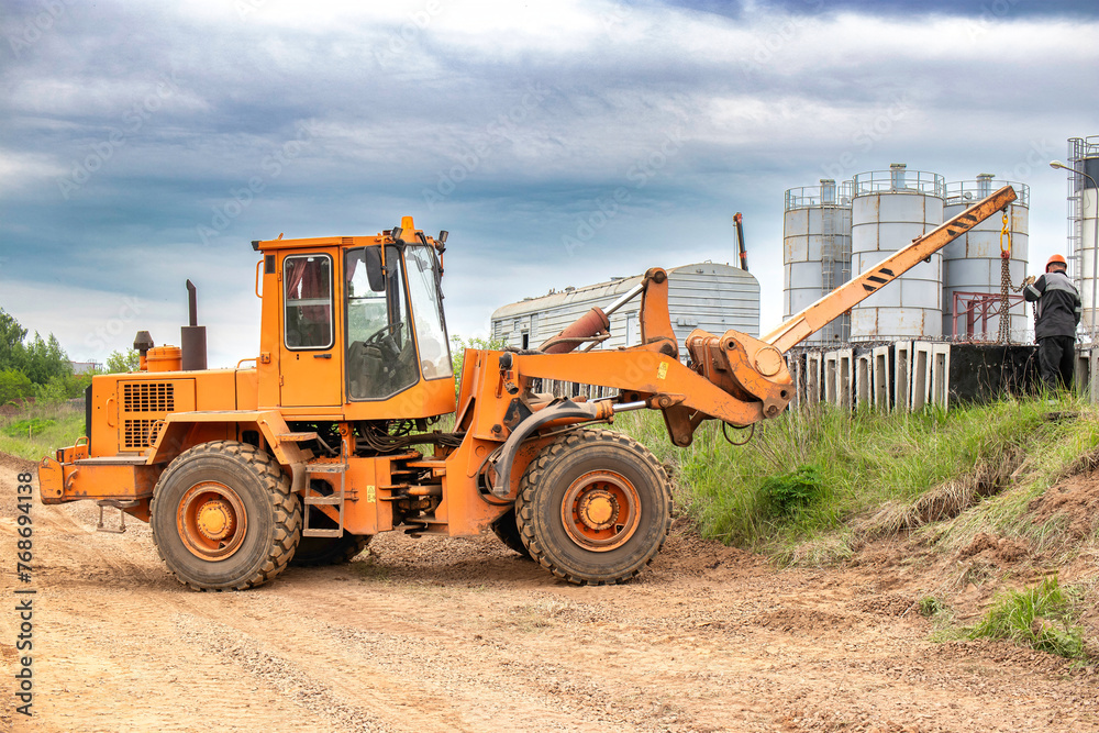 A tractor or forklift is stationary on the side of a dusty rural road, surrounded by fields and under a clear sky A tractor or forklift is stationary on the side of a dusty rural road, surrounded by fields and under a clear sky