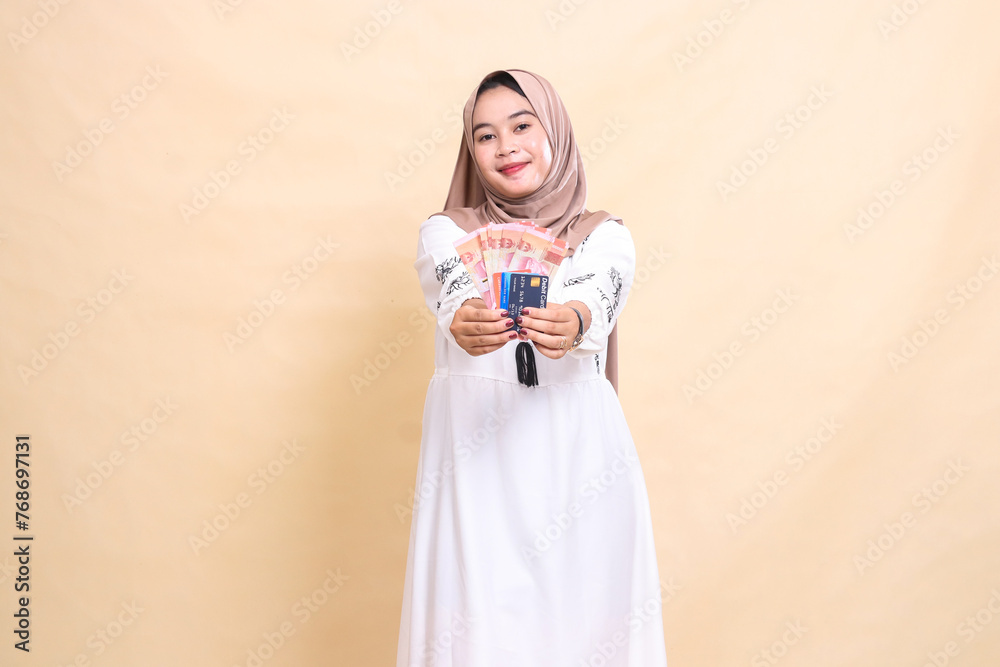 A beautiful indonesia Muslim woman wearing a hijab smiles and brings forward rupiah notes and a debit credit card for payment. used for advertising, technology, Eid and Ramadan A beautiful indonesia Muslim woman wearing a hijab smiles and brings forward rupiah notes and a debit credit card for payment. used for advertising, technology, Eid and Ramadan