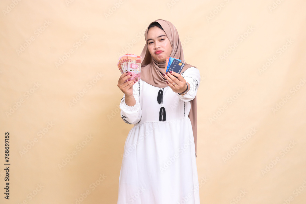 a beautiful Asian Muslim girl in a hijab sullenly carries rupiah money and advances a debit credit card for payment. used for advertising, technology, Eid and Ramadan a beautiful Asian Muslim girl in a hijab sullenly carries rupiah money and advances a debit credit card for payment. used for advertising, technology, Eid and Ramadan