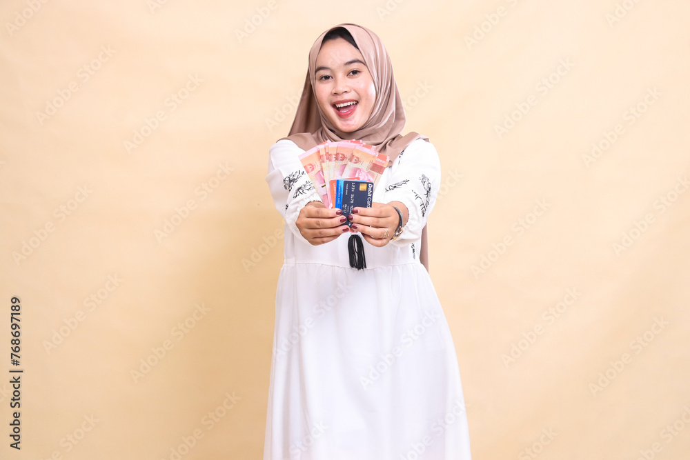 a beautiful Asian Muslim woman wearing a happy hijab brings forward rupiah money and a debit credit card for payment. used for advertising, technology, Eid and Ramadan a beautiful Asian Muslim woman wearing a happy hijab brings forward rupiah money and a debit credit card for payment. used for advertising, technology, Eid and Ramadan