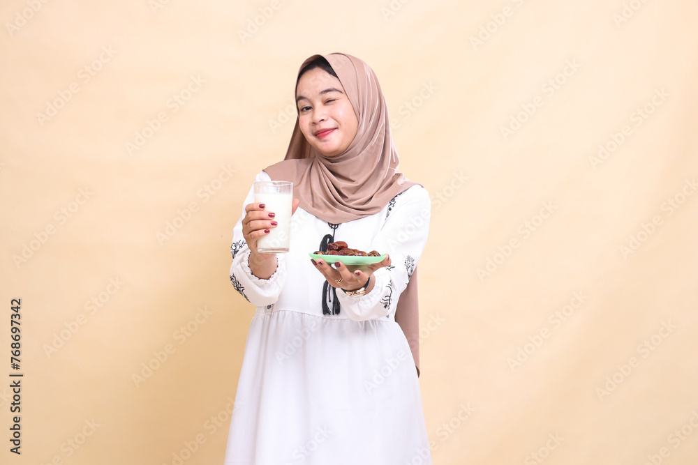 A beautiful Asian Muslim woman wearing a hijab smiles with a wink and brings a glass of milk and dates to the front while breaking the fast. used for advertising, health, Eid and Ramadan A beautiful Asian Muslim woman wearing a hijab smiles with a wink and brings a glass of milk and dates to the front while breaking the fast. used for advertising, health, Eid and Ramadan