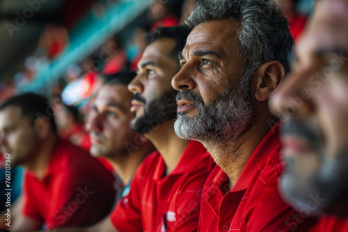 A man in a red shirt is sitting in a stadium watching a game. A group of men wearing red shirts are sitting in a stadium, watching a game. A group of men wearing red shirts are sitting in a stadium