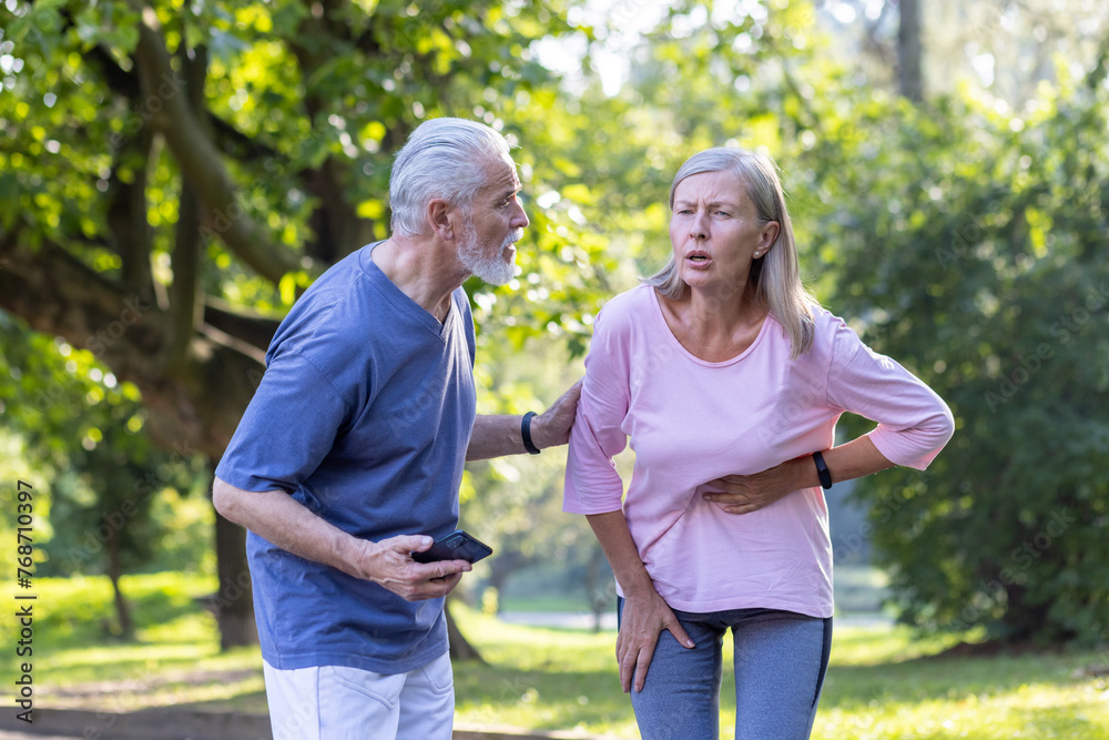 Senior couple doing sports and walking in the park, the woman feels severe pain in the side of the body and in the heart, the man tries to help, calls an ambulance on the phone. Senior couple doing sports and walking in the park, the woman feels severe pain in the side of the body and in the heart, the man tries to help, calls an ambulance on the phone.