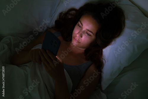 Late at night, a woman rests in bed, drawn to her phone's glow,an image emblematic of insomnia and social media's hold. 
