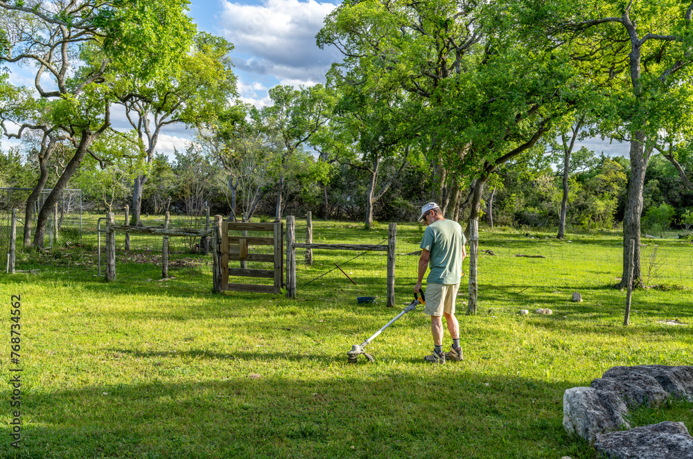 Matured Caucasian man using an electric battery operated grass line trimmer in an area with trees, a fence and a gate Matured Caucasian man using an electric battery operated grass line trimmer in an area with trees, a fence and a gate