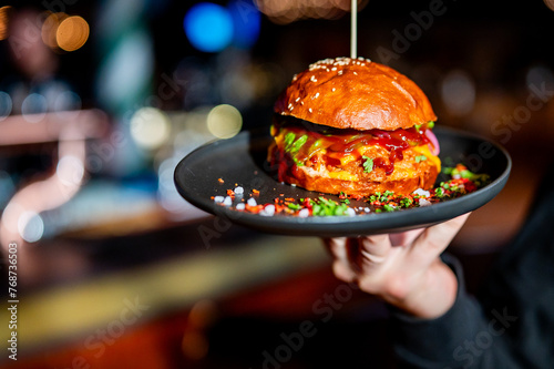 A hand holds a black plate with a juicy burger, garnished with lettuce and sauce, on a sesame seed bun. The blurred background suggests a bar or restaurant setting.