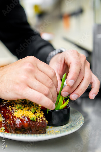 close-up view person, likely a chef, garnishing prepared dish. The hands are in focus, placing green herbs onto the food. The dish appears to be a meaty entree with golden-brown crumbs or seasoning. 