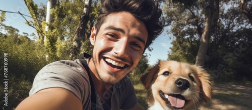 A young man is smiling and holding up his phone to take a selfie with his dog, who is also looking at the camera. They are outside in a park, enjoying each others company and the fresh air.