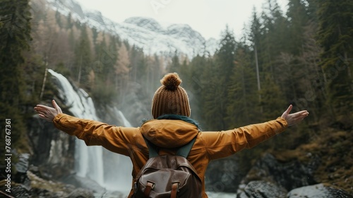 Person in yellow jacket with open arms facing a majestic waterfall in a forested mountain landscape.
