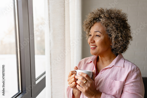 A mature woman of mixed race looking outside a large window, holding a cup of coffee and smiling