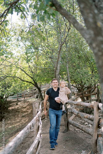 Dad with a little girl in his arms walks along a path between fences in the park