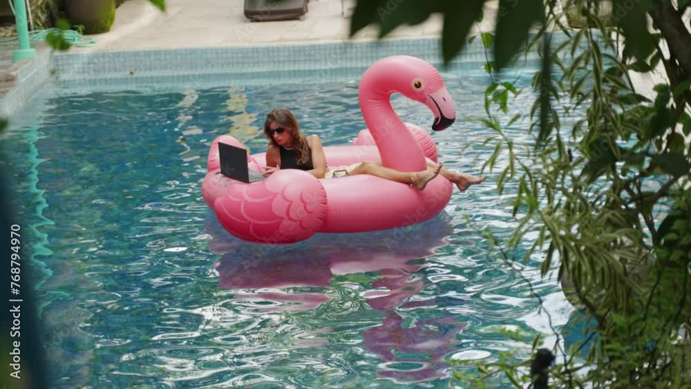 Young woman lying on a pink flamingo inflatable at swimming pool and working with laptop