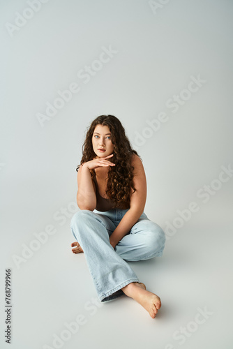 charming curvy woman in 20s with curly brown hair sitting and looking to camera with hand to chin