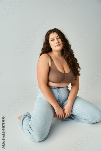 pretty plus size young woman in brown bra and blue jeans sitting against grey background