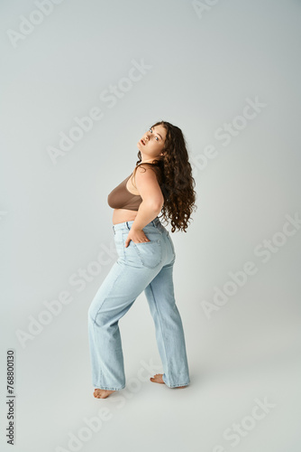 seductive plus size young woman in brown bra and blue jeans putting foot forward on grey background