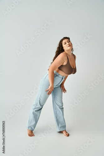 beautiful plus size woman in brown bra and blue jeans leaning to forward on grey background