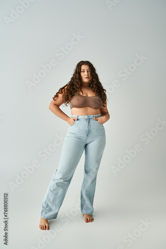 plus size woman in brown bra and blue jeans with curly hair posing against grey background