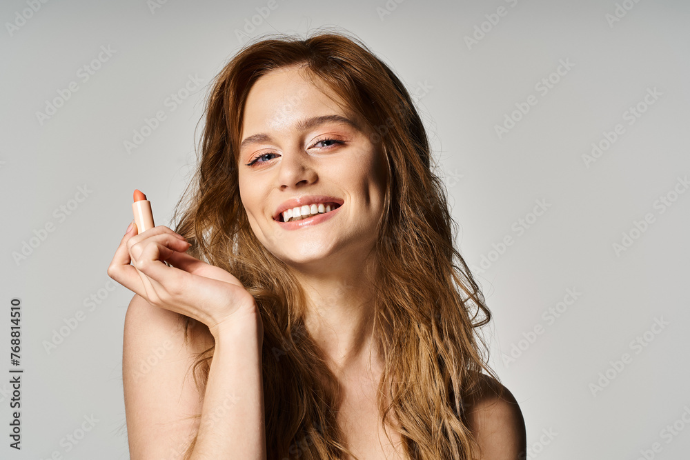 Beauty shot of smiling woman looking at camera with nude makeup holding lipstick on grey background Beauty shot of smiling woman looking at camera with nude makeup holding lipstick on grey background