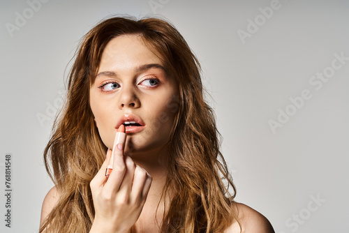 Beauty portrait of woman looking away with nude makeup applying lipstick on grey background