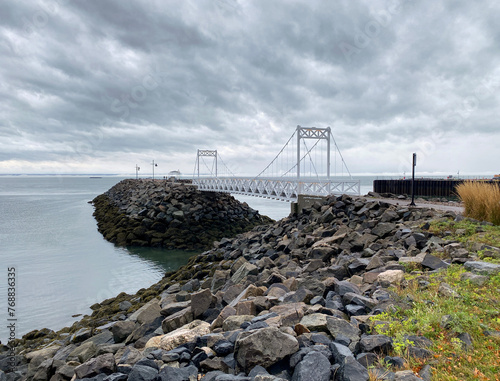 Pier on the coast. Structure of a pedestrian bridge on a quay. Walkway at the river's edge on a cloudy day. White gate on a rocky bank. Boat and buoy in the distance.
