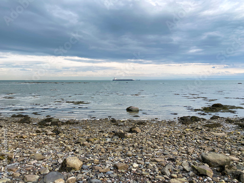 Beach and rocks on a cloudy day. Coastal beach at low tide. Stones on the coast. Cargo boat in the distance on the river.