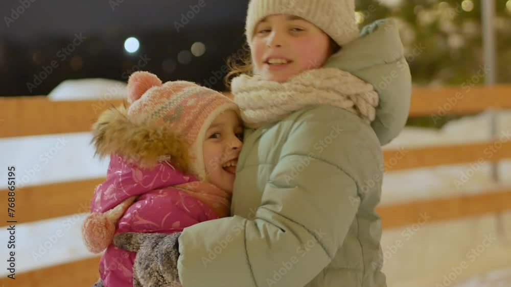 Two little girls sisters on the street in winter at the ice rink in warm clothes against the background of street Christmas lights smiling hugging looking into the frame. Love in the family. Two little girls sisters on the street in winter at the ice rink in warm clothes against the background of street Christmas lights smiling hugging looking into the frame. Love in the family.