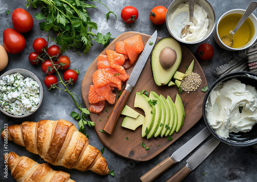 Croissants, salmon pieces, avocado pieces, knife, pot with cream cheese on the cutting board flat lay, ingredients for breakfast concept