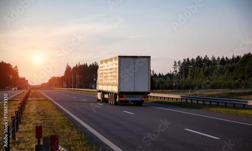 A truck with a tilt semi-trailer carries cargo along the highway in the evening against the backdrop of a summer sunset. The concept of cargo transportation, the logistics, the work and rest  © HENADZY