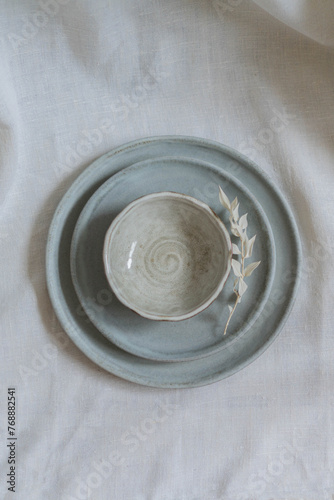 A spool of white thread perched atop a set of hand-thrown ceramic plates. The plates are stacked with a graceful symmetry, their cool grey tones complementing the crisp white of both thread and linen.
