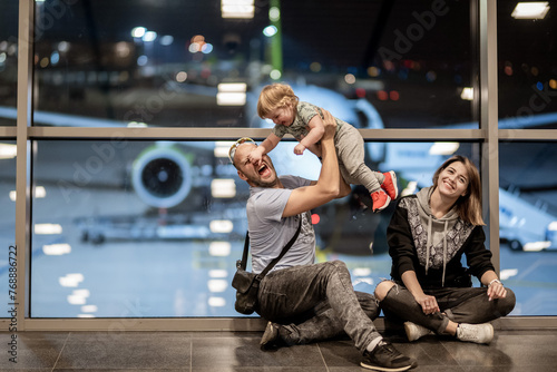 Riga, Latvia - October 19, 2019 - A joyful family moment at the airport: a man playfully holds a toddler above him while sitting next to a smiling woman, with an airplane in the backdrop