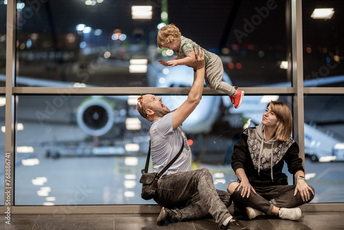 Riga, Latvia - October 19, 2019 - A man lifts a laughing toddler into the air while sitting beside a woman at an airport, with a plane visible through the window behind them.