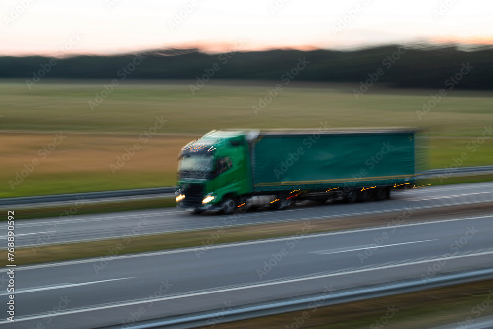 A modern truck with a semi-trailer and headlights on transports cargo in the evening, in the summer, against the backdrop of a field. Trade turnover between countries. Import and export. Blurred