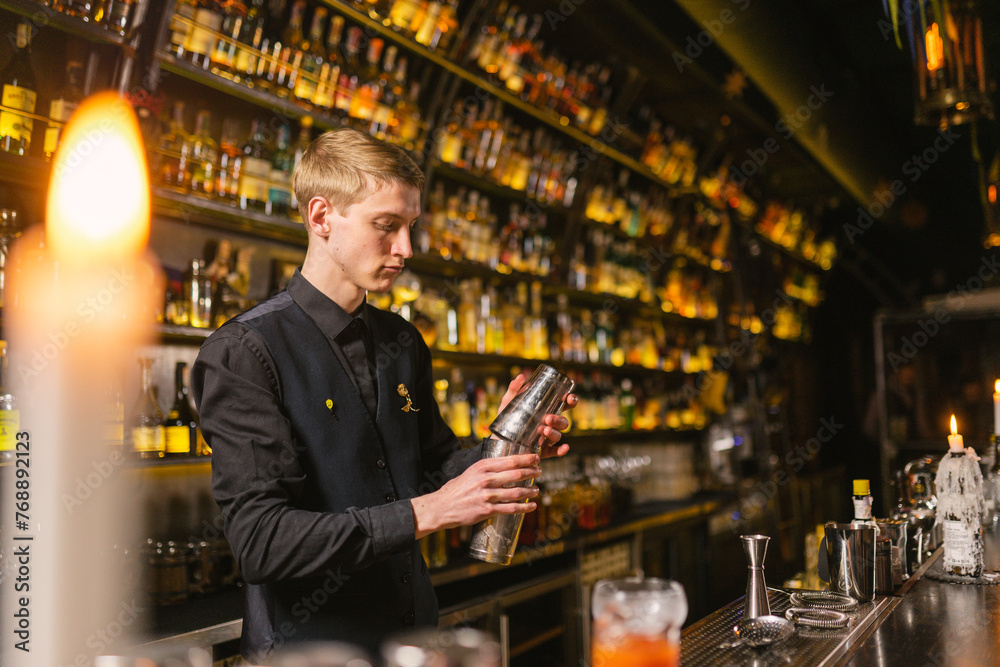 Concentrated barkeeper makes shakes for guests of pub using bar metal shaker. Young barman works in antique-style bar with huge selection of alcohol