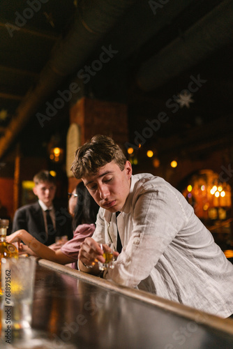 Guy in depressed state sits at bar and drinks alcohol from shot glass. Young guy sits at wooden bar counter with glass of elite cognac