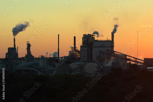 Large factory structure with smoke from production process rising up polluting atmosphere at manufacturing yard. Industrial site at sunset