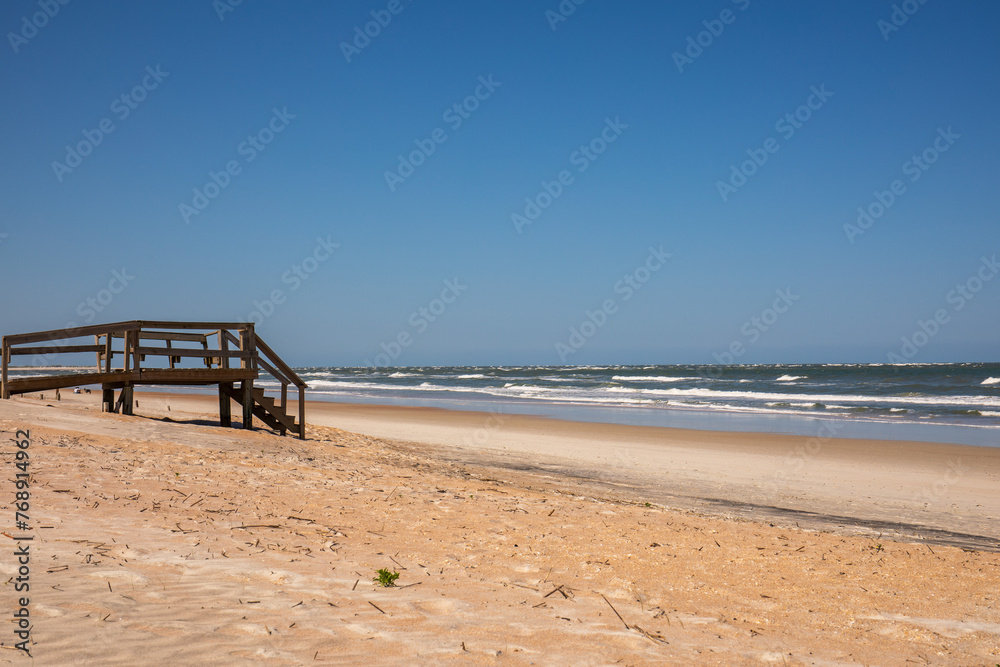Sandy shore on Florida beach.  Small waves and sandy beach in the foreground with blue sky and water.  Sand dunes in the foreground with wooden plank stairs and boardwalk.  Blue sky and white sand.