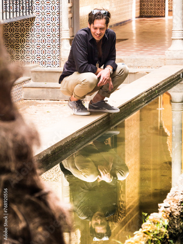 Thirty-year-old man looks smiling at himself in the reflection of a fountain while crouching