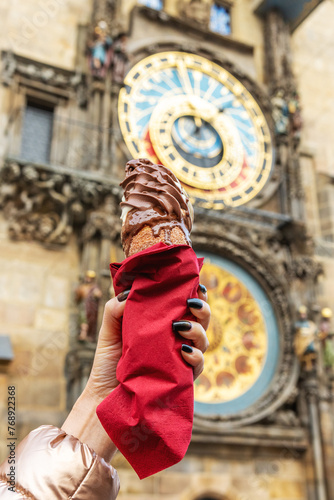 Photography Female hand is holding traditional czech cookie trdelnik on background of Astronomical clock in Old Town of Prague city