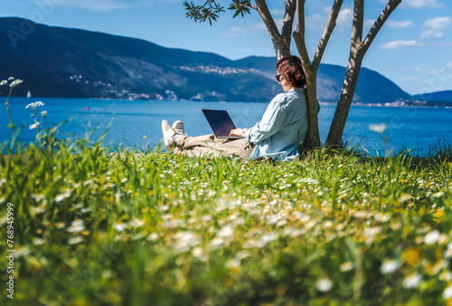 Young woman sitting on a green summer lawn on the seashore using a laptop