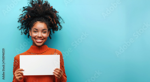 A young smiling Black woman in an orange sweater holds up a white blank paper sheet on a blue background, banner for advertising copy space, blank sign for your advertising text messages