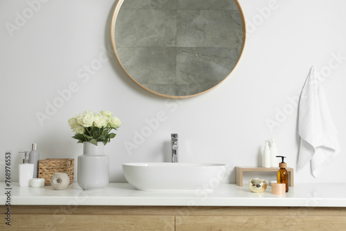 Vase with beautiful white roses and toiletries near sink in bathroom