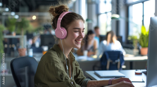 young woman is wearing pink headphones and smiling while working on a computer in a busy office environment