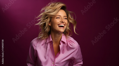 A radiant woman with blonde hair laughs wholeheartedly against a deep purple backdrop, wearing a stylish pink shirt photo