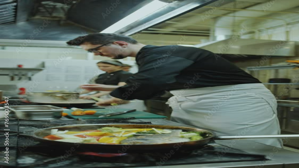 Chef adding leek and cherry tomatoes to fish with vegetables in cooking pan on stove while preparing food at work in restaurant kitchen. Vertical clip Chef adding leek and cherry tomatoes to fish with vegetables in cooking pan on stove while preparing food at work in restaurant kitchen. Vertical clip