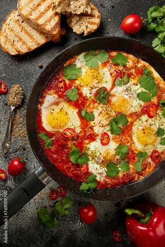 Tasty Shakshuka eggs in a pan with toast on a dark concrete background. Eggs poached  in a spicy tomato pepper sauce. Traditional Jewish scrambled eggs. Flat lay, Top view.