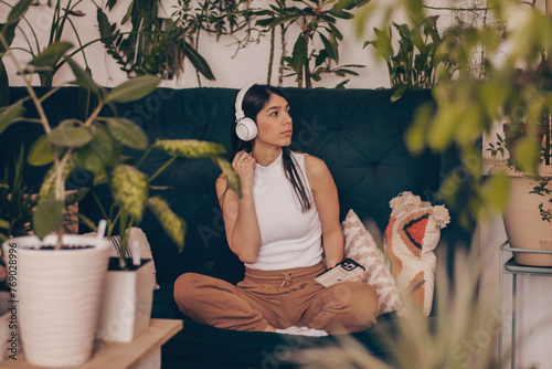 A young dark-haired woman in casual clothes wearing headphones,using a smartphone,sitting on the couch.Relaxation,meditation,listening music.