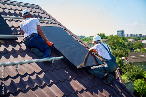 Workers building solar panel system on rooftop of house. Two men installers in helmets installing photovoltaic solar module outdoors. Alternative, green and renewable energy generation concept.