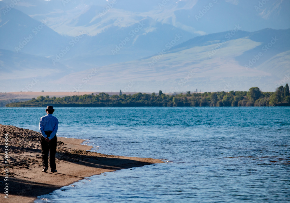 An old man walking by the shore of the famous Issyk-Kul lake surrounded by the Ala-Too mountains. Kyrgyzstan, July 2023