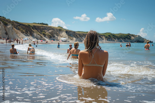 Rear view of a woman sitting in the sea, facing towards sunlit cliffs and other beach goers photo