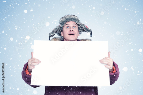 A happy man in winter clothes holds a white empty banner in his hands and looks at the sky with his mouth open. On a light blue background with snow